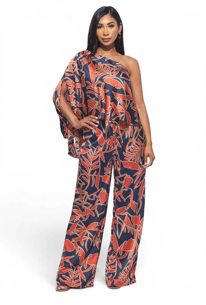 Woman wearing a tropical satin one-shoulder top with draped sleeve and matching wide leg pants featuring a coral and navy botanical print.